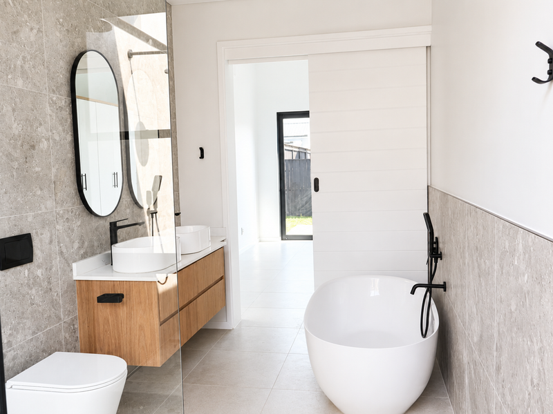 Modern bathroom featuring a double vanity, freestanding bathtub, wall-mounted toilet, and contemporary black matte fixtures with neutral stone-look wall tiling.