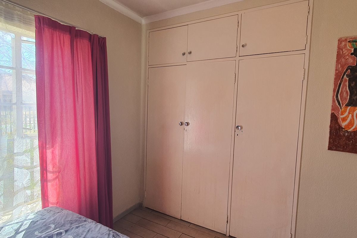 A bedroom featuring built-in cupboards, tiled flooring, and a window with security bars, offering a functional space with potential for cosmetic updates.