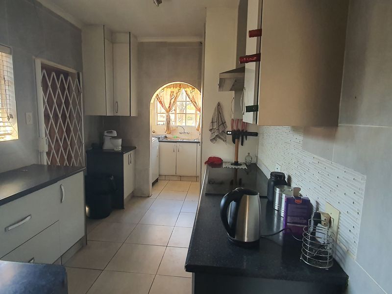 A functional kitchen featuring built-in cabinetry, dark laminate countertops, tiled backsplash, and ceramic flooring, with an adjoining scullery area visible through the archway.