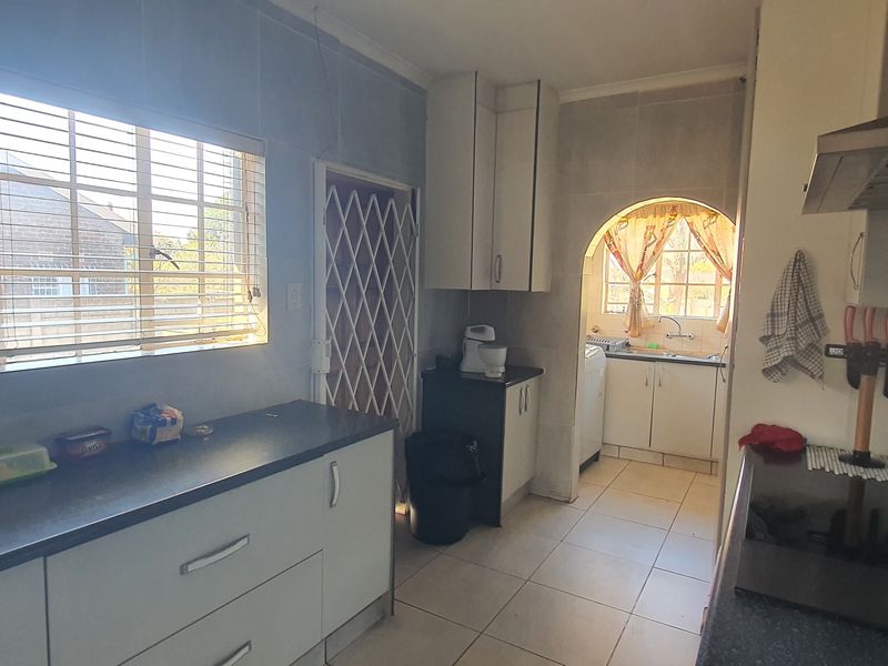A functional kitchen featuring built-in cabinetry, dark laminate countertops, and a tiled splashback, with an adjoining scullery area visible through an arched doorway.