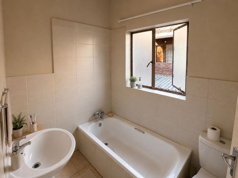 A functional bathroom featuring a bathtub, pedestal basin, and toilet with neutral wall tiling and a window providing natural light.