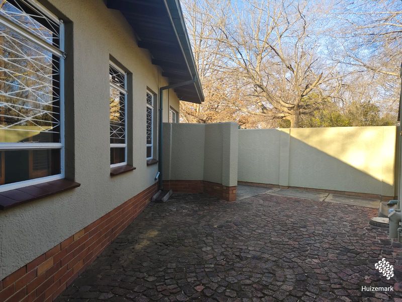 A rear exterior view of the property featuring a cobblestone paved courtyard, boundary wall, and security-fitted windows.