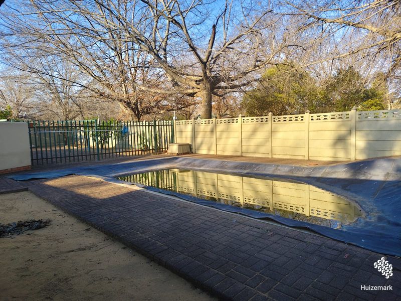 Outdoor pool area featuring a swimming pool with a cover, surrounded by paved decking and enclosed by a precast concrete wall and metal fencing.