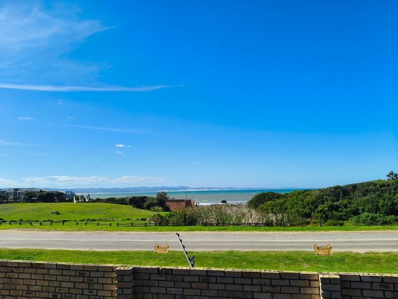 A scenic coastal view overlooking a green verge, road, and distant ocean horizon, framed by a brick boundary wall with electric fencing.
