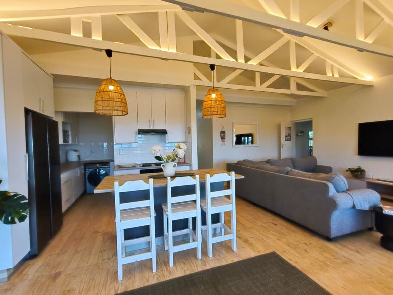 Bright open-plan living area featuring a modern kitchen with subway tile backsplash, exposed timber trusses, and a central island, seamlessly flowing into the lounge.