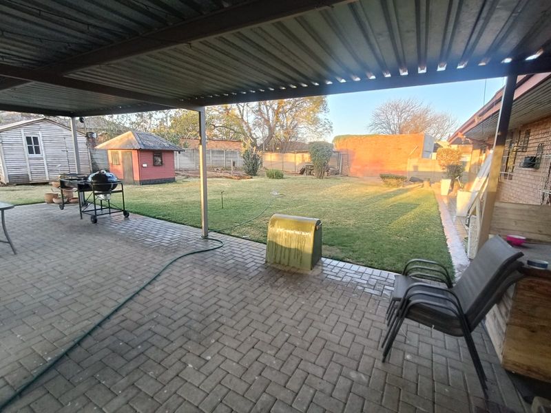A covered paved patio area overlooking a spacious backyard garden with a lawn and outbuildings.