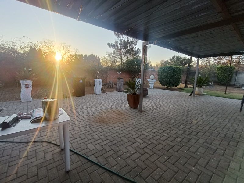 A spacious covered patio area featuring paved flooring and a metal roof structure, overlooking a private garden with a brick boundary wall.
