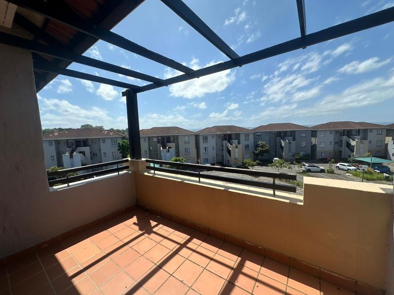 An open-air balcony featuring terracotta tiling, a metal pergola structure, and a metal balustrade, offering views of the surrounding apartment complex.