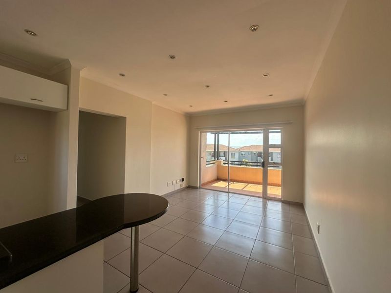 An open-plan living area featuring neutral tiled flooring, recessed lighting, and a breakfast counter, with direct access to an outdoor balcony.