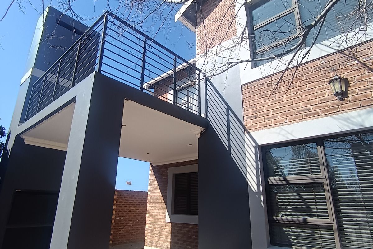 Rear view of a modern townhouse featuring a brick facade, a covered patio with a built-in braai, and an upper-level balcony with metal railings.