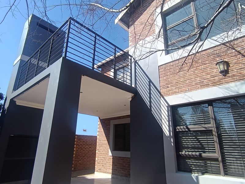 Rear view of a modern townhouse featuring a brick facade, a covered patio with a built-in braai, and an upper-level balcony with metal railings.
