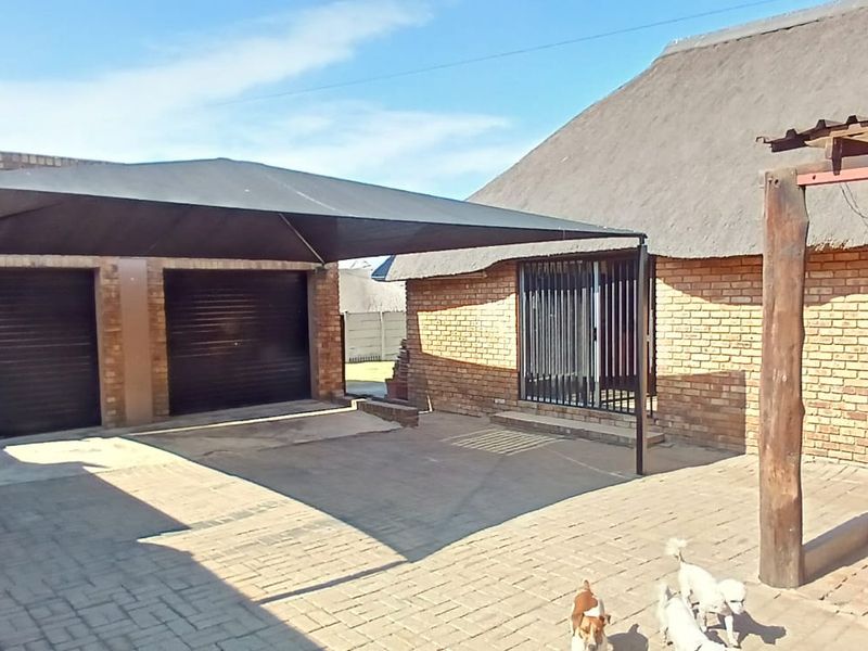 Front exterior view featuring a double garage with roller doors, a shade sail carport, and a brick-built structure with a traditional thatch roof.