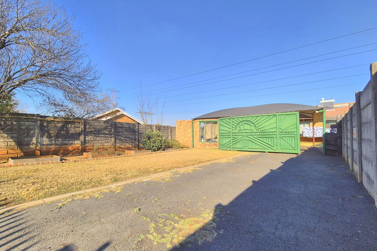 Rear exterior view featuring a paved driveway, a large metal sliding gate, a carport, and a precast concrete boundary wall enclosing the yard.