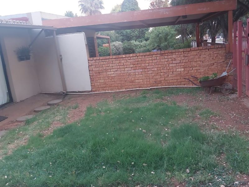 A rear exterior view featuring a small lawn area, a brick boundary wall, a carport structure, and a sliding glass door entrance.