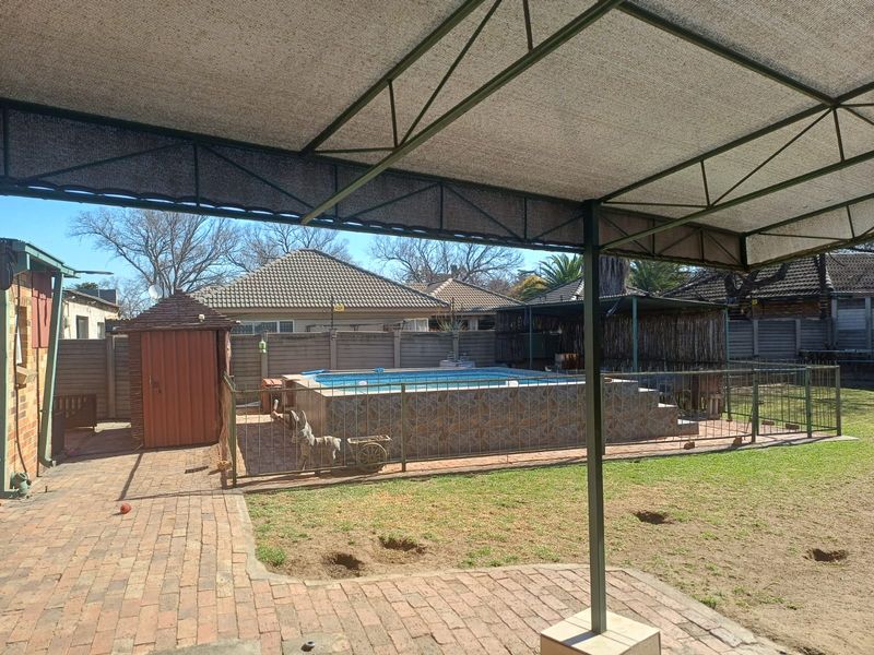 Outdoor backyard area featuring a raised swimming pool with tiled surround, enclosed by metal fencing, set within a paved and lawned garden space.