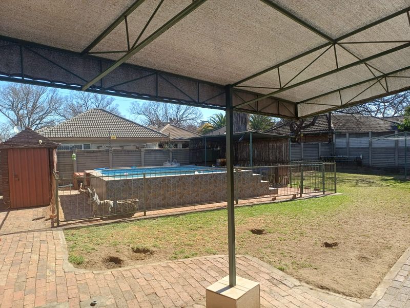 Outdoor pool area featuring an above-ground swimming pool with tiled surround, enclosed by metal fencing, set within a garden with brick paving and shade cover.