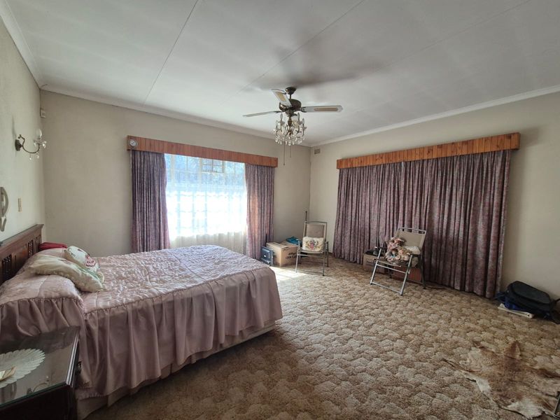 Spacious bedroom featuring a ceiling fan, crystal chandelier, and timber curtain pelmets, offering a dated interior ready for modernization.
