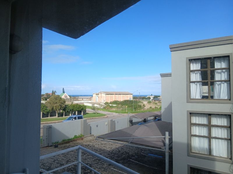 An elevated view from a balcony overlooking the apartment complex grounds, paved parking area, and surrounding neighborhood towards the distant coastline.