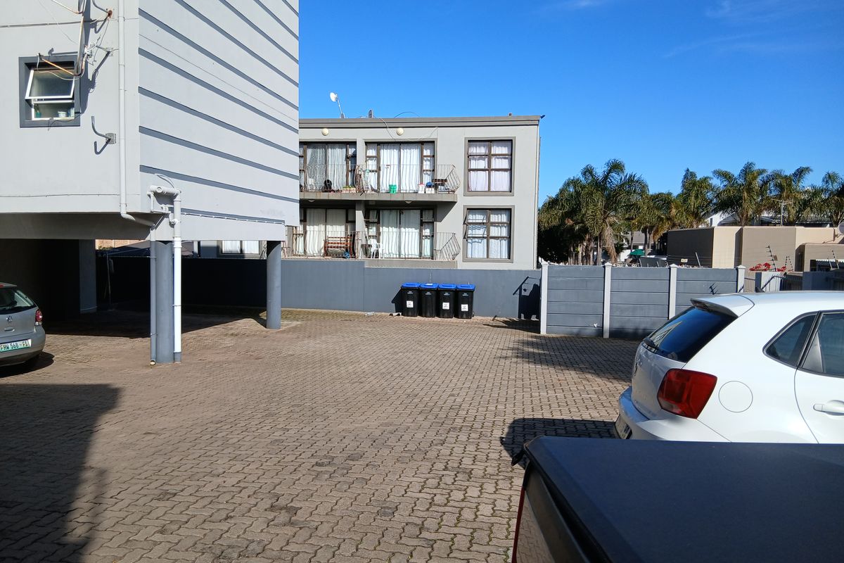 Rear exterior view of the apartment complex featuring a paved parking area, boundary wall, and residential balconies.