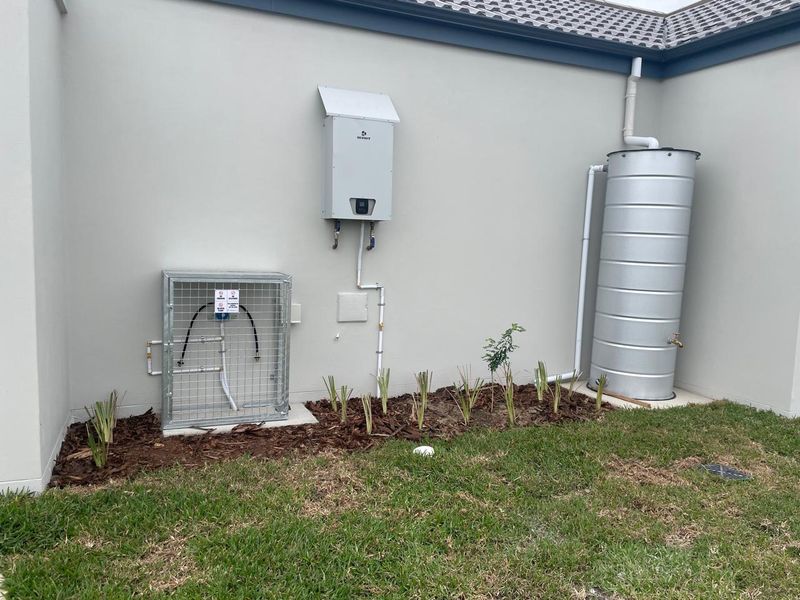 Exterior view showing a wall-mounted gas water heater, a metal rainwater tank with downpipe, and a protective cage for utility connections.