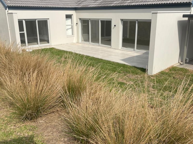 Rear view of the property featuring a paved patio area, large sliding glass doors, and a well-maintained lawn with landscaped garden beds.