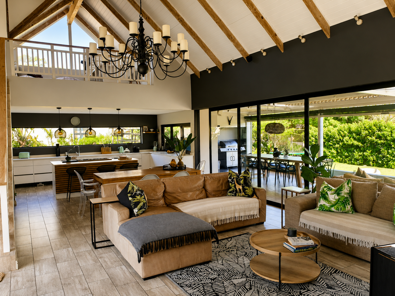 Spacious open-plan living and kitchen area featuring a vaulted timber ceiling, large chandelier, and seamless indoor-outdoor flow through expansive sliding glass doors.