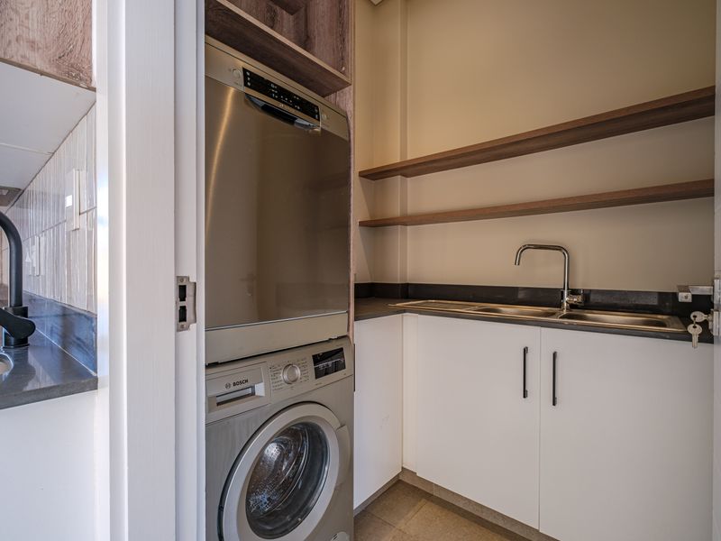 A compact, modern scullery featuring built-in cabinetry, stone countertops, a stainless steel sink, and integrated laundry appliances.