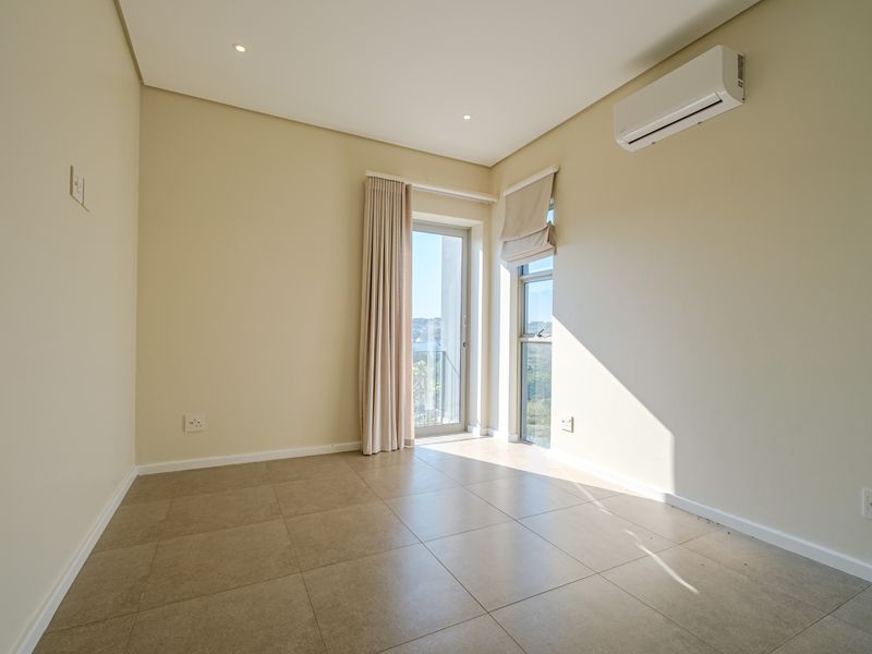 A bright, empty bedroom featuring neutral tiled flooring, recessed lighting, a wall-mounted air conditioning unit, and access to a balcony through a glass door.