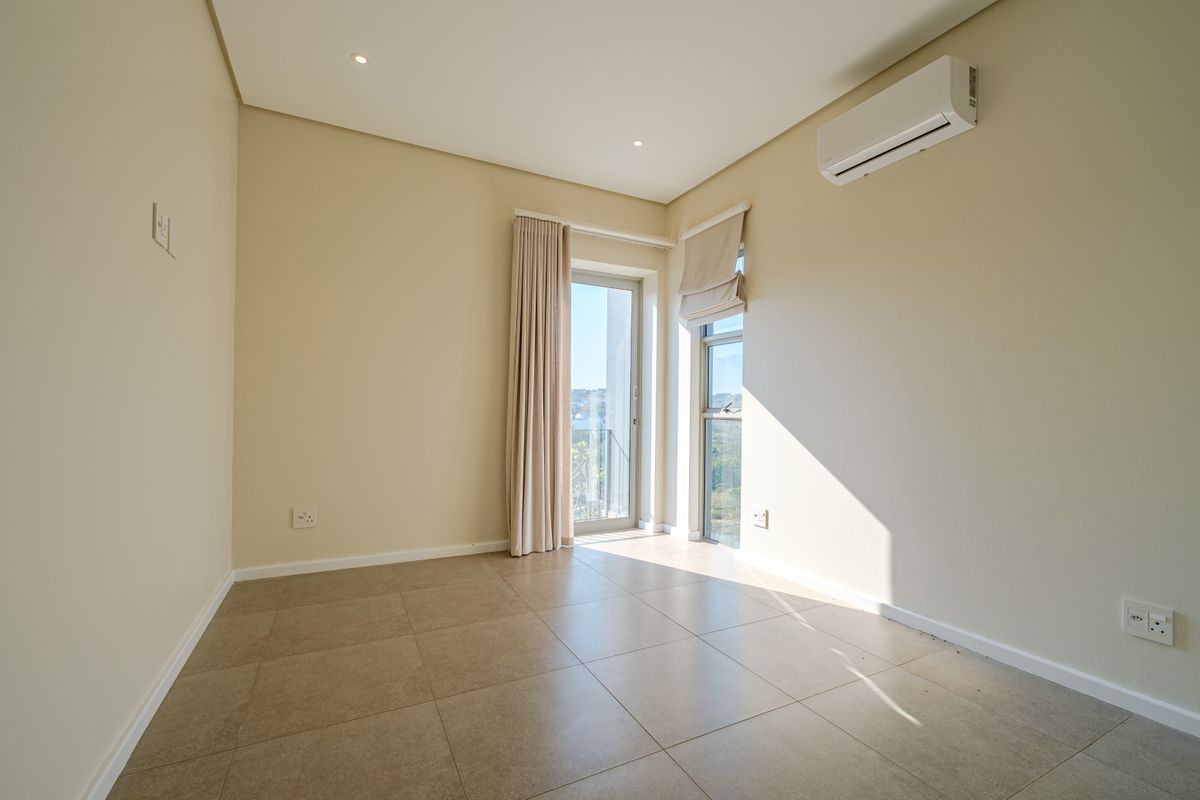 A bright, empty bedroom featuring neutral tiled flooring, recessed lighting, a wall-mounted air conditioning unit, and access to a balcony through a glass door.