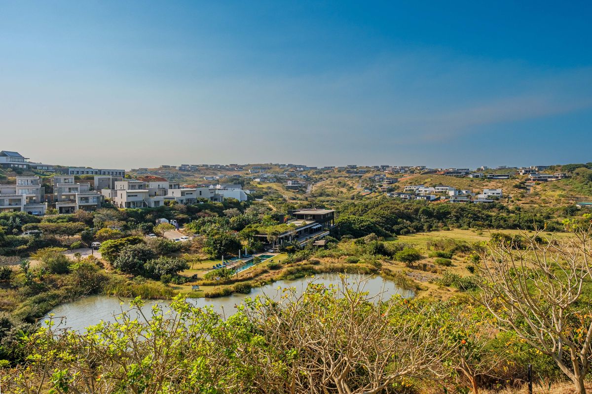 An elevated aerial view of a residential estate featuring a natural pond, lush greenery, and modern apartment buildings set within a scenic landscape.