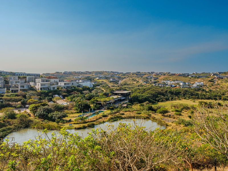 An elevated aerial view of a residential estate featuring a natural pond, lush greenery, and modern apartment buildings set within a scenic landscape.