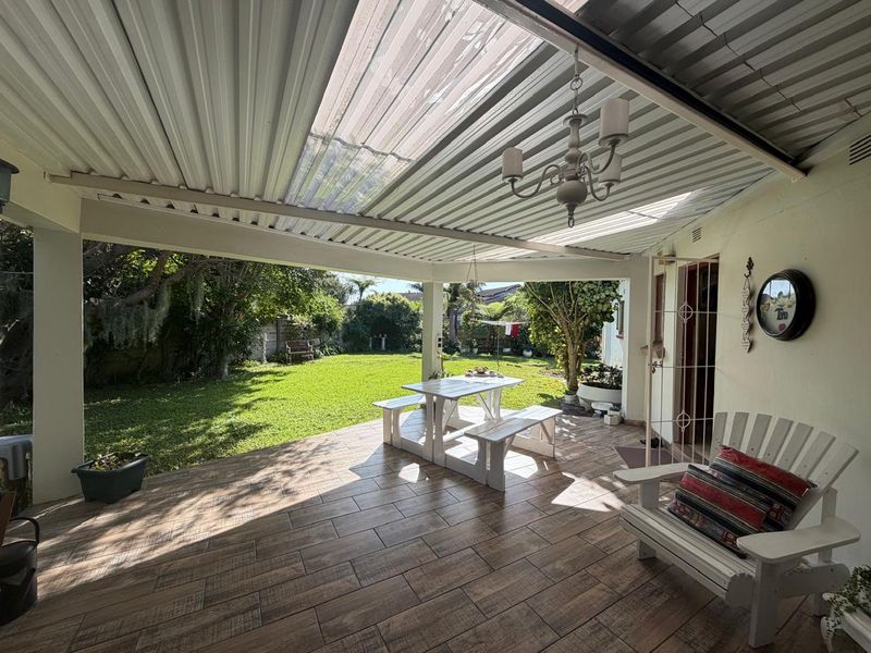 A covered outdoor patio featuring tiled flooring, a corrugated roof with translucent panels, and direct access to a spacious garden area.