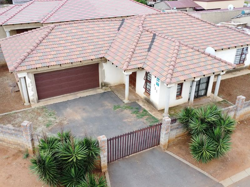 Aerial view of a single-story residential house featuring a tiled roof, attached garage, paved driveway, and perimeter walling with a metal gate.
