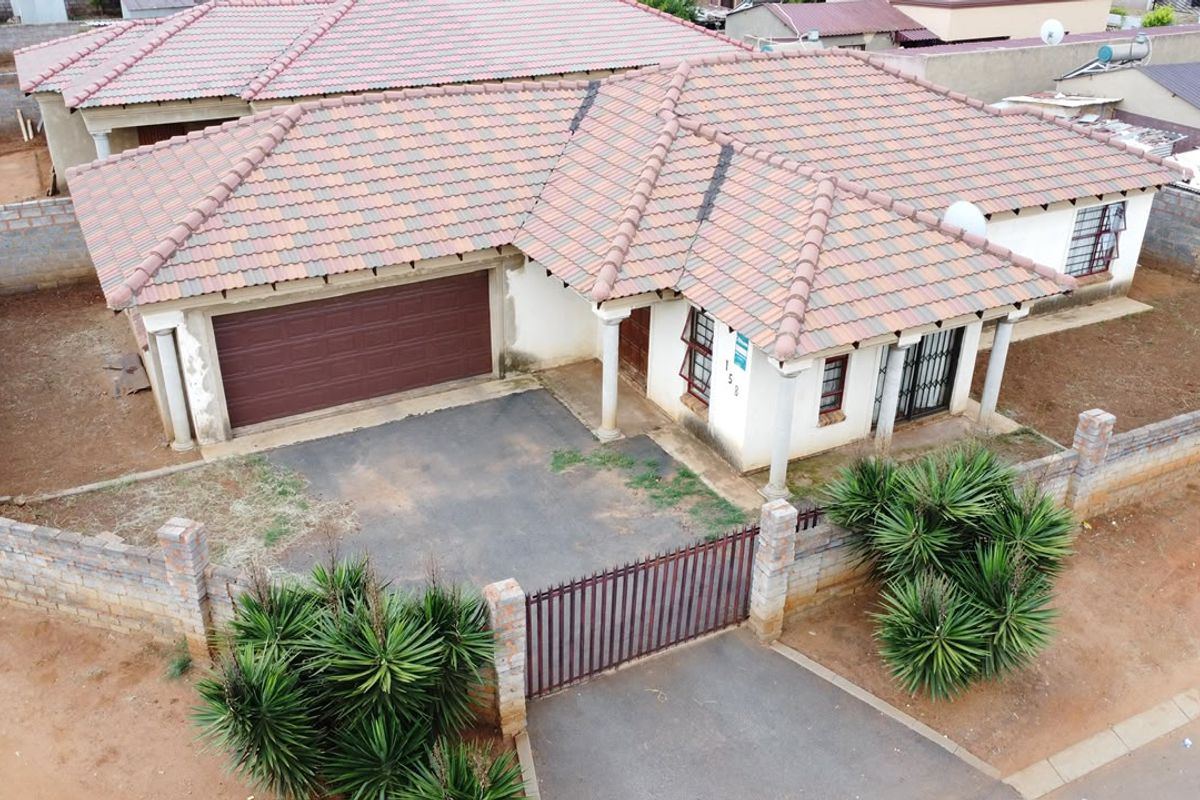 Aerial view of a single-story residential house featuring a tiled roof, attached garage, paved driveway, and perimeter walling with a metal gate.