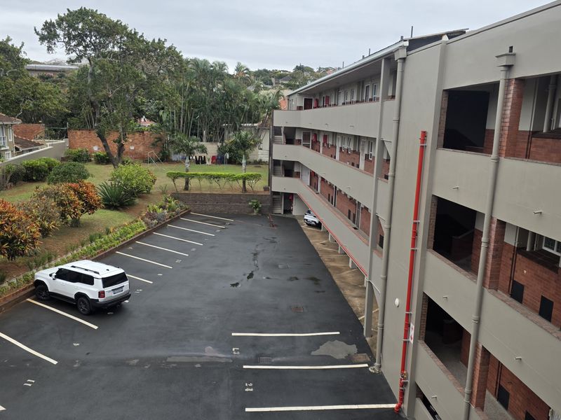 An elevated view of the apartment complex rear exterior, featuring a paved parking area, landscaped garden, and multi-level building facade.