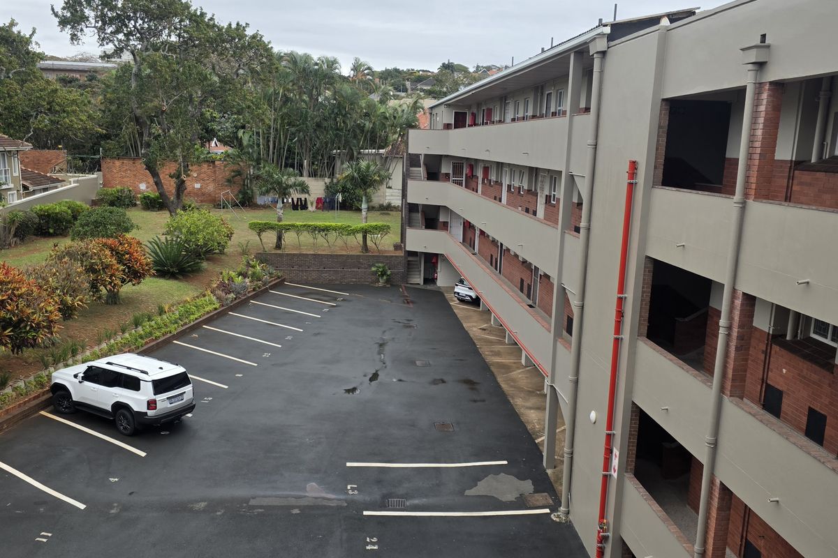 An elevated view of the apartment complex rear exterior, featuring a paved parking area, landscaped garden, and multi-level building facade.
