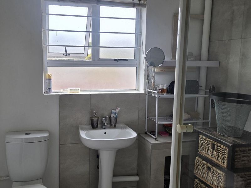 A functional bathroom featuring a pedestal washbasin, toilet, and a glass-enclosed shower with tiled finishes and natural light from a top-hung window.