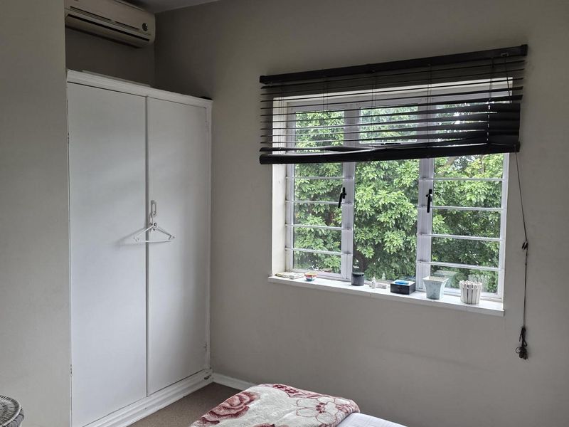 A bedroom featuring a built-in wardrobe, a wall-mounted air conditioning unit, and a window with security bars and venetian blinds.