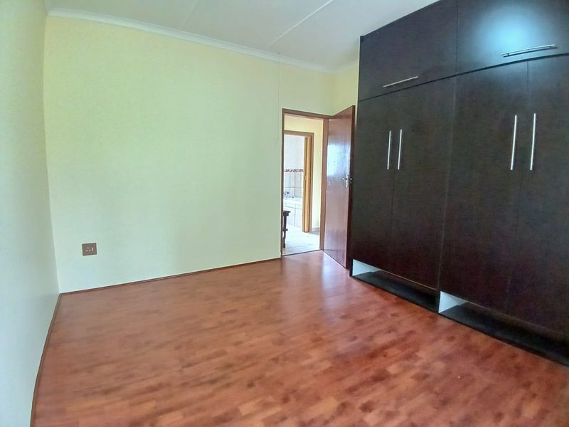 A spacious, empty bedroom featuring dark wood-finish built-in cupboards and warm-toned laminate flooring, with a doorway leading to an adjacent bathroom.
