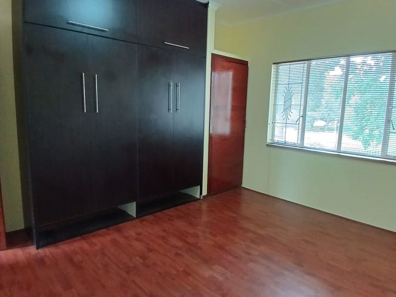 A clean, empty bedroom featuring dark wood-finish built-in cupboards, warm-toned laminate flooring, and a window with security bars.