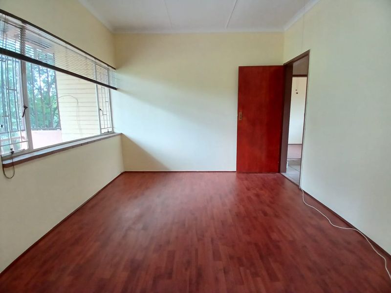 A bright, empty bedroom featuring laminate flooring, a large window with venetian blinds, and a solid timber door.