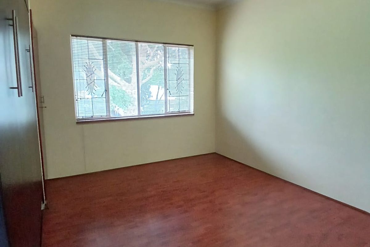 A clean, empty bedroom featuring laminate flooring, built-in cupboards, and a window fitted with security bars and venetian blinds.