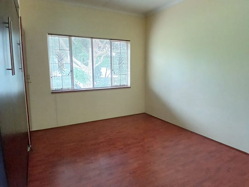 A clean, empty bedroom featuring laminate flooring, built-in cupboards, and a window fitted with security bars and venetian blinds.