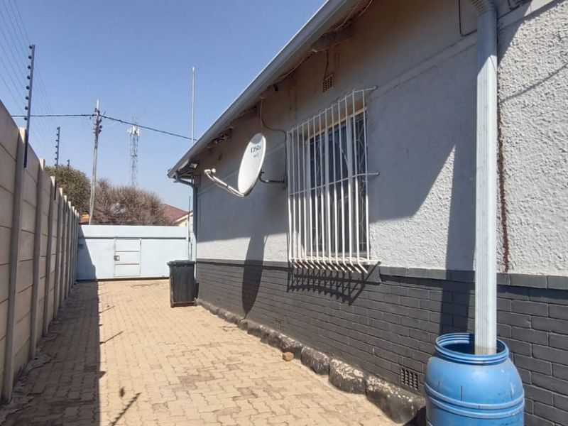 A side exterior view of the property featuring a paved walkway, precast boundary wall with electric fencing, and security bars on the window.