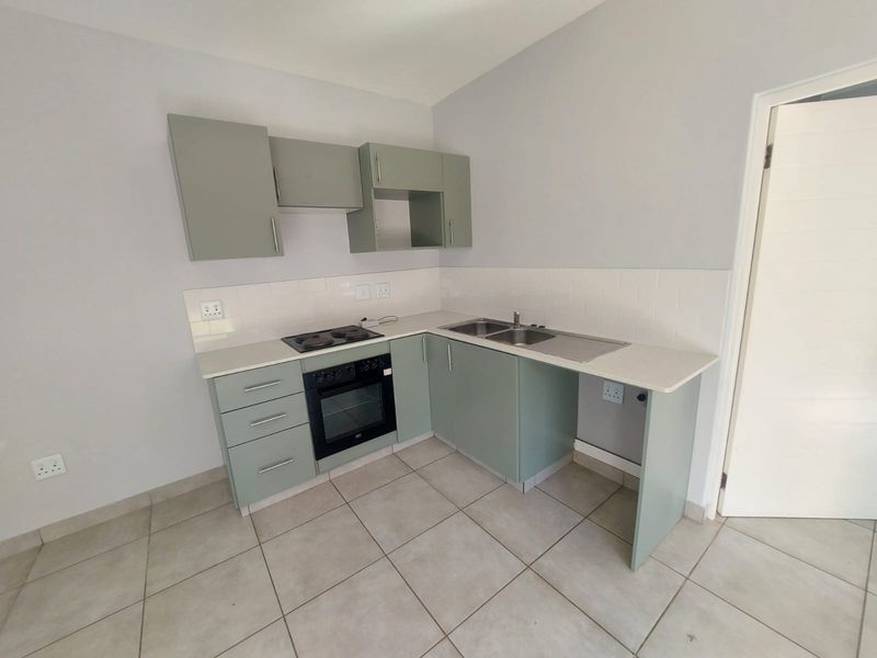A clean, modern kitchen featuring sage green cabinetry, a built-in oven and hob, stainless steel sink, and neutral tiled flooring.
