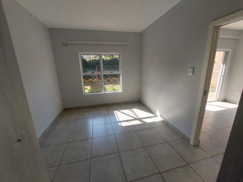 A bright, empty bedroom featuring neutral tiled flooring and a window overlooking the garden area.