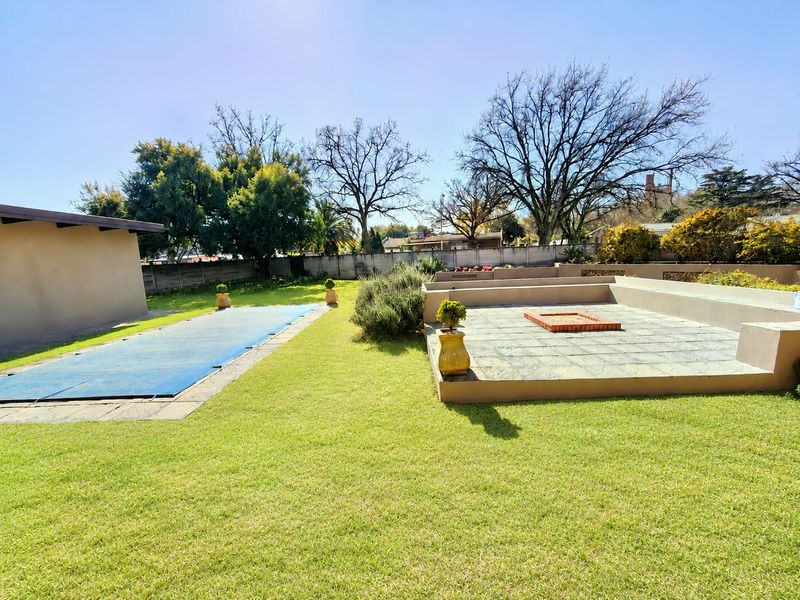 Spacious backyard featuring a swimming pool with cover, a raised paved patio area with a built-in brick fire pit, and established lawn.