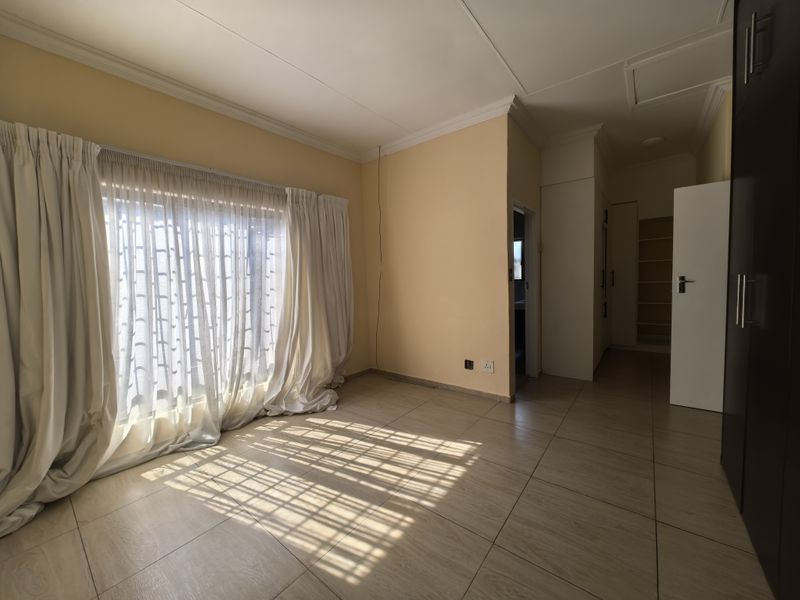 A spacious, empty bedroom featuring neutral tiled flooring, built-in cupboards, and large windows with curtains, offering a clean and versatile living space.