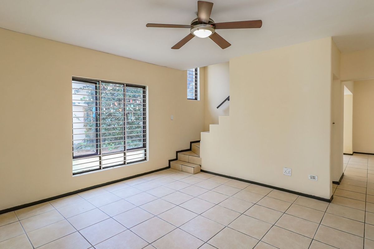 An empty living room featuring tiled flooring, a ceiling fan, and an internal staircase with security bars on the windows.