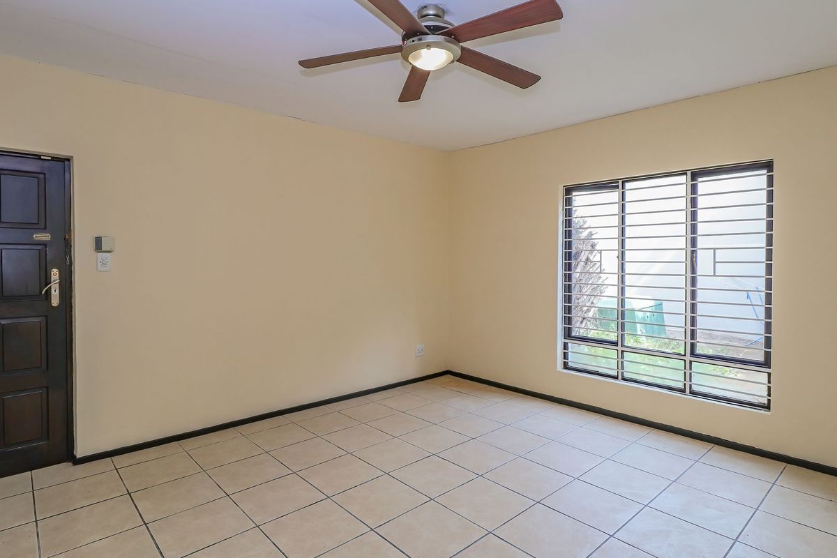 A spacious, empty bedroom featuring neutral tiled flooring, a ceiling fan, and a secure window with fitted burglar bars.